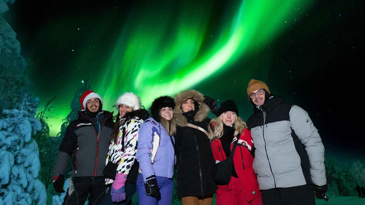 Travellers in thermal overalls under a green and violet aurora on a small-group aurora borealis tour Rovaniemi departs for each evening, Finland